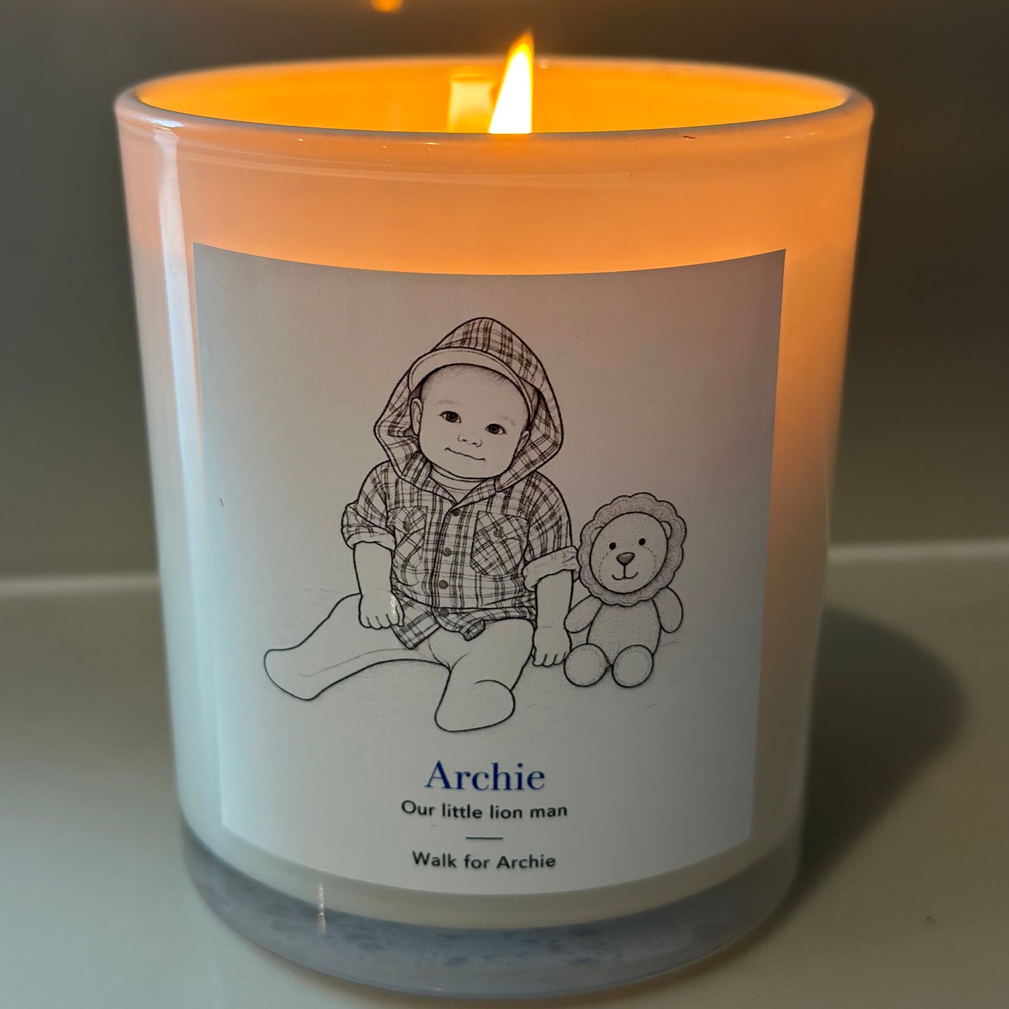 A lit white glass candle jar with a warm orange glow, featuring a custom printed label with a baby illustration and the name 'Archie'.